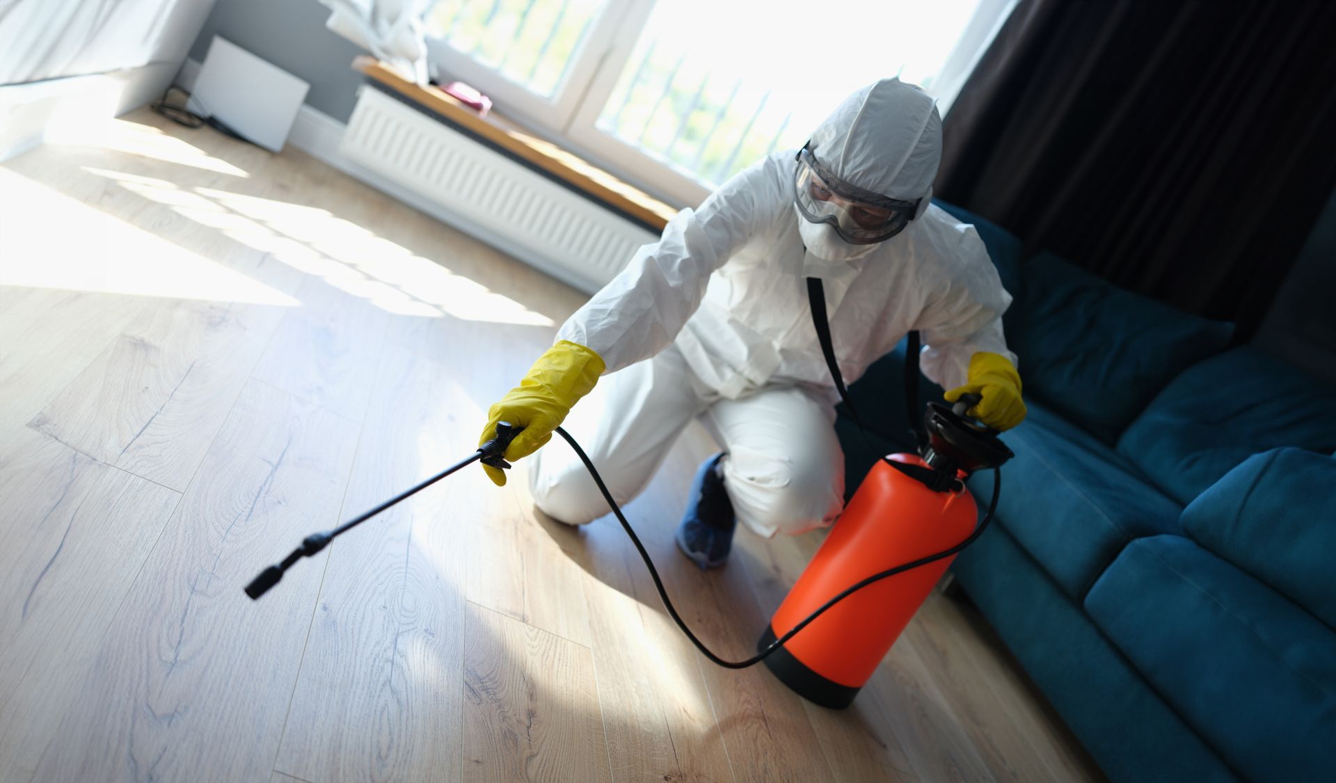 Person in protective suit spraying floor with a red and black pesticide tank in a living room.