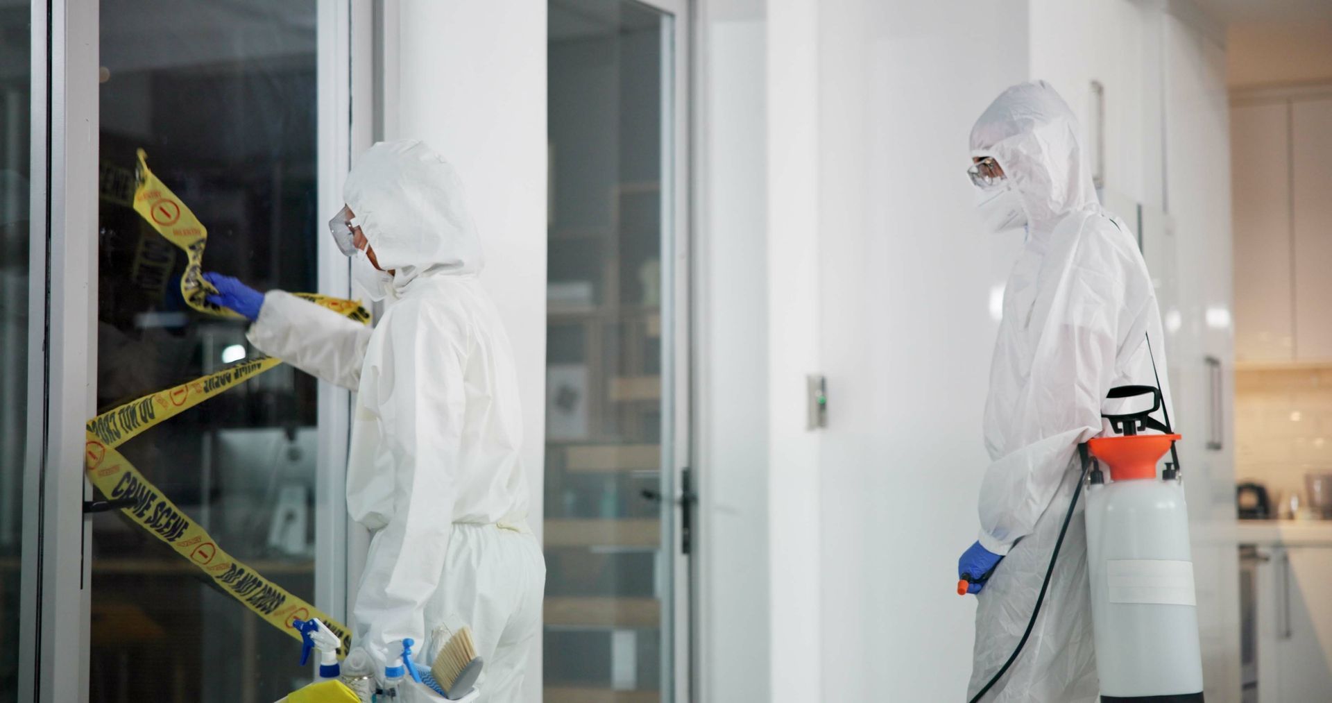 Two people in hazmat suits investigate a scene, one taping caution tape, the other with a sprayer.