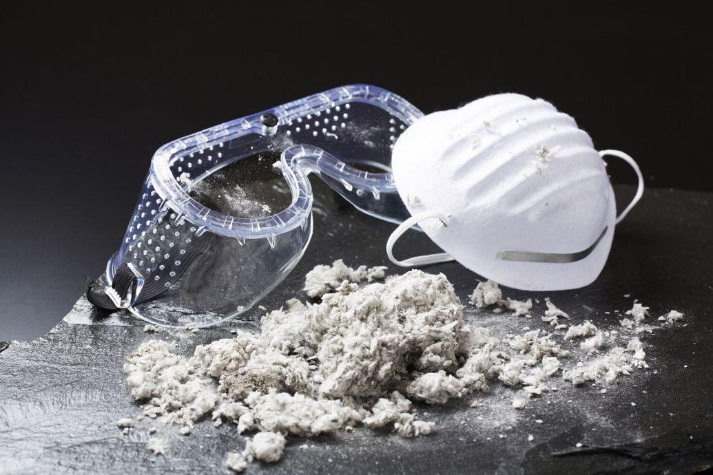 Protective eyewear and mask next to a pile of gray asbestos fibers on a dark surface.