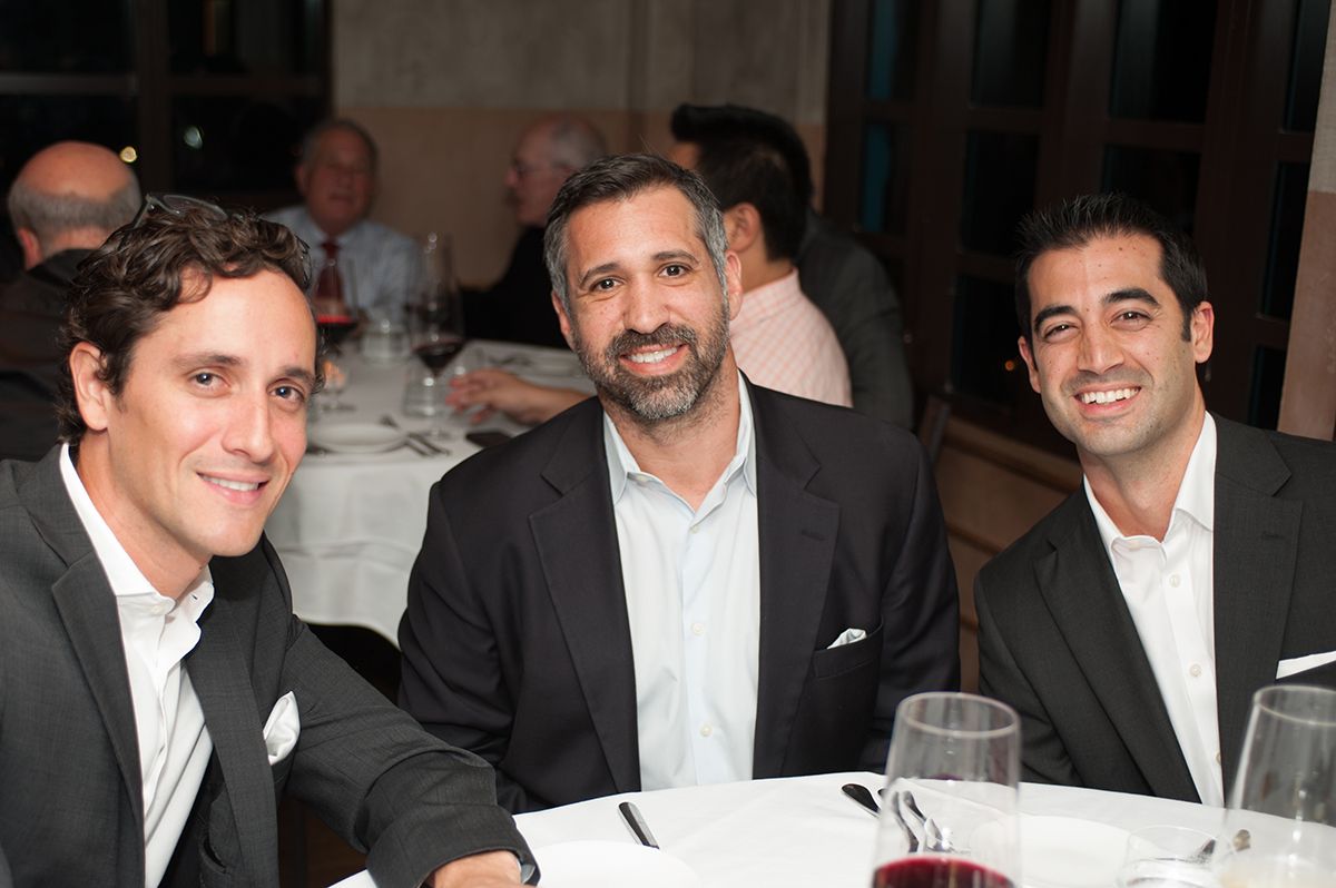 Three men are posing for a picture at a table with wine glasses.