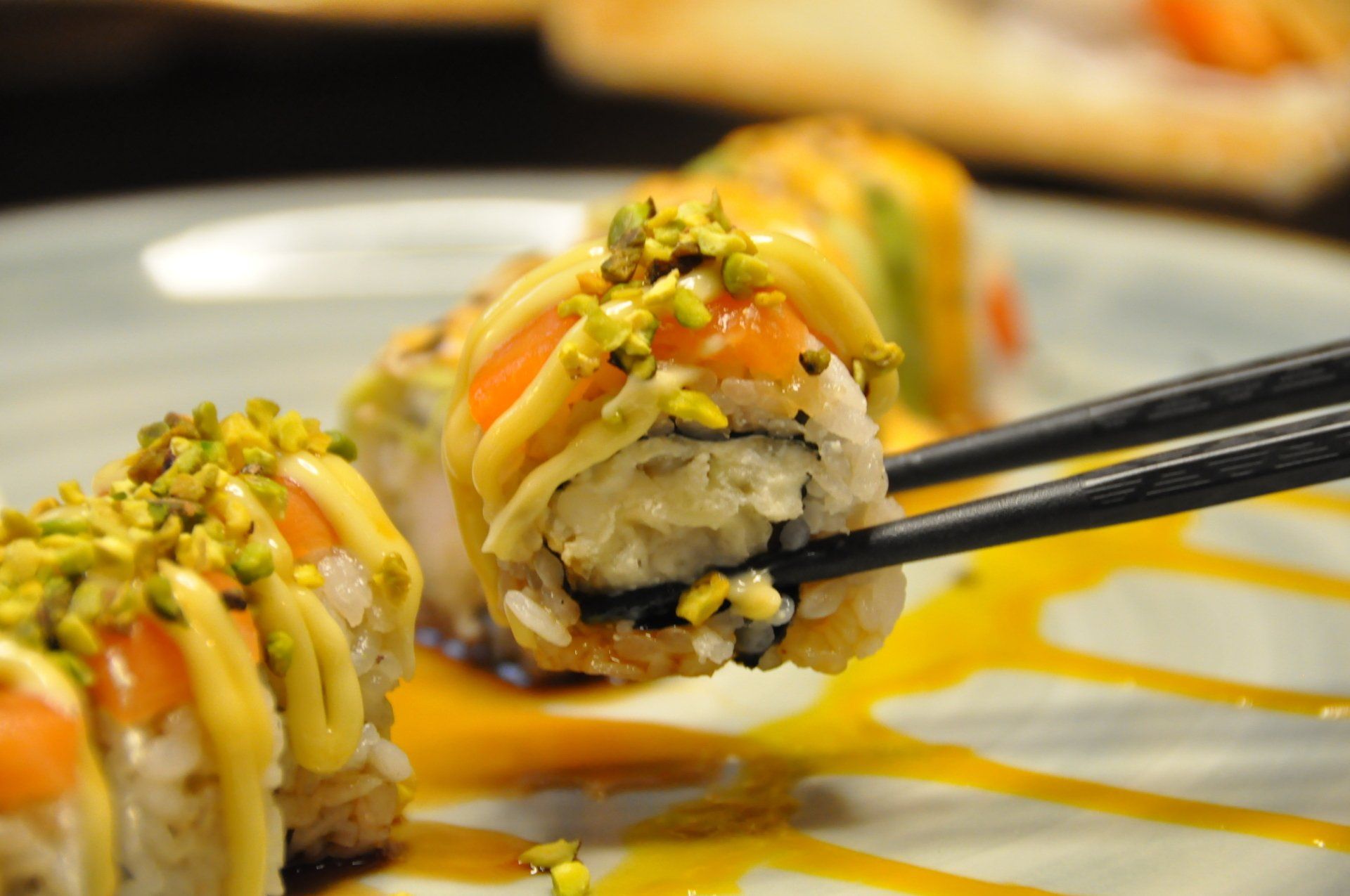 Sushi roll servito con bacchette su un piatto bianco, irrorato di salsa e guarnito con frutta secca tritata.