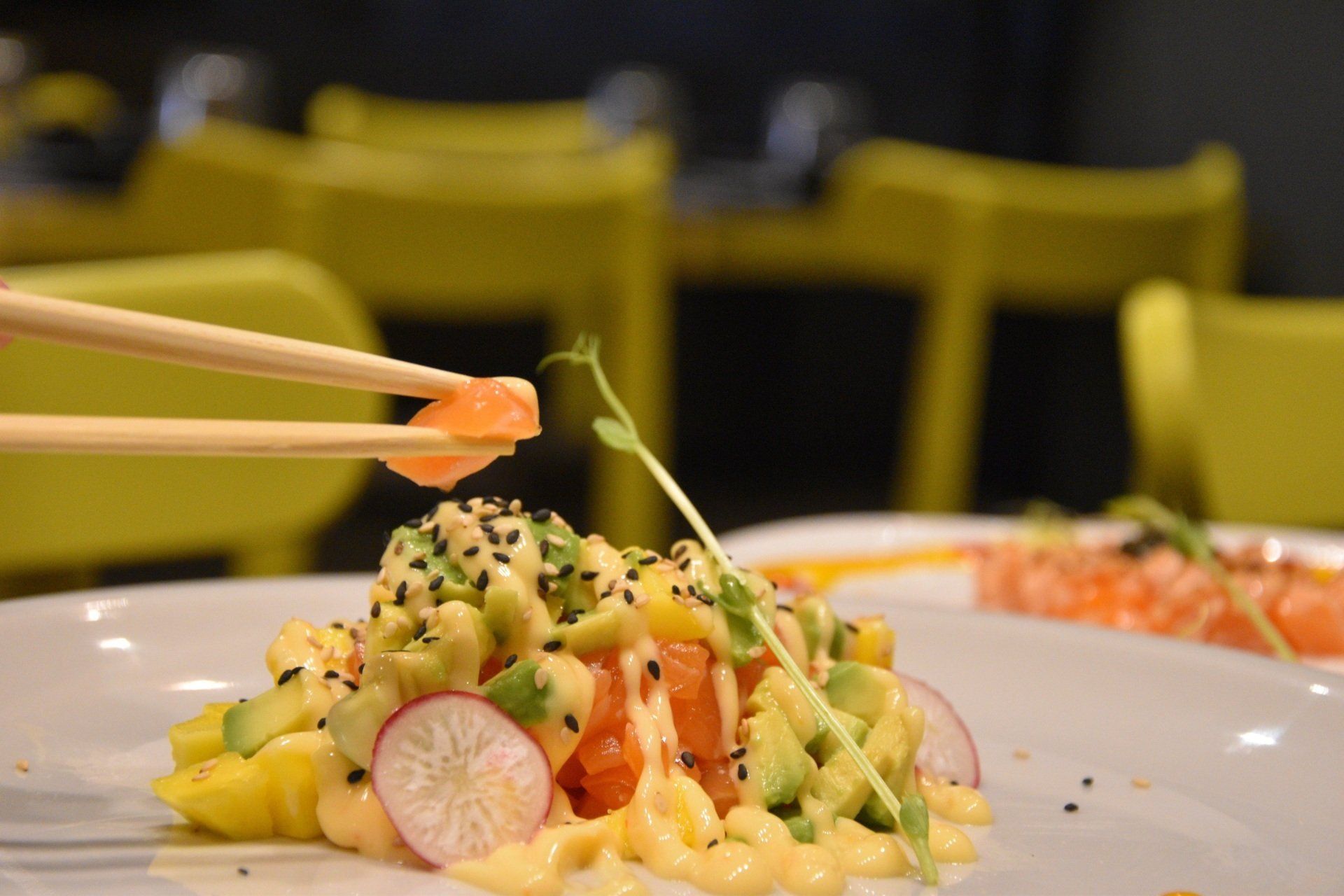 Bacchette che sollevano rotolini di sushi su un piatto bianco, guarniti con ravanello ed erbe aromatiche, in un ambiente da ristorante.