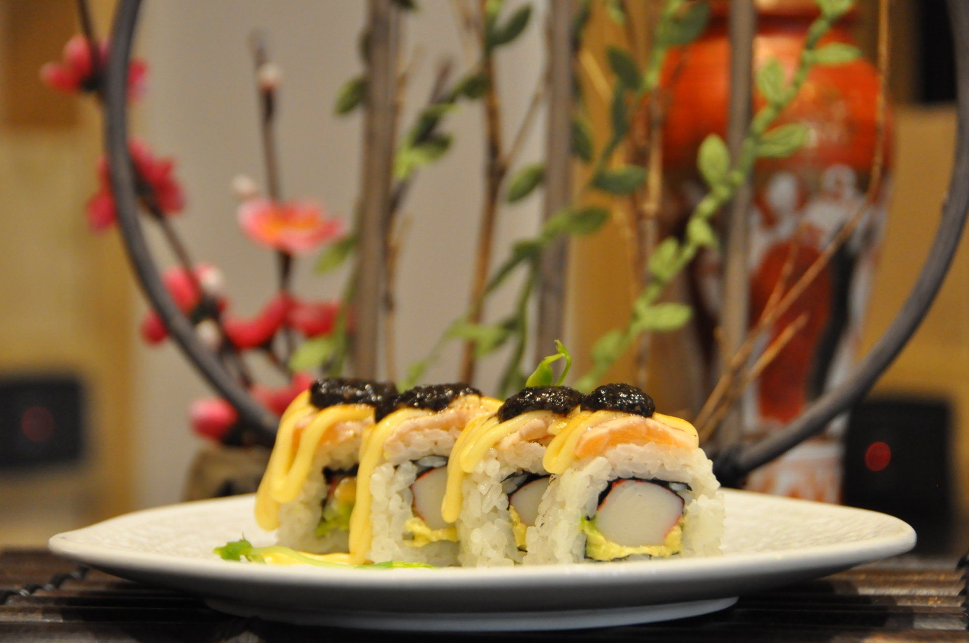 Rotoli di sushi su un piatto bianco con salsa di soia e fiori decorativi sullo sfondo.