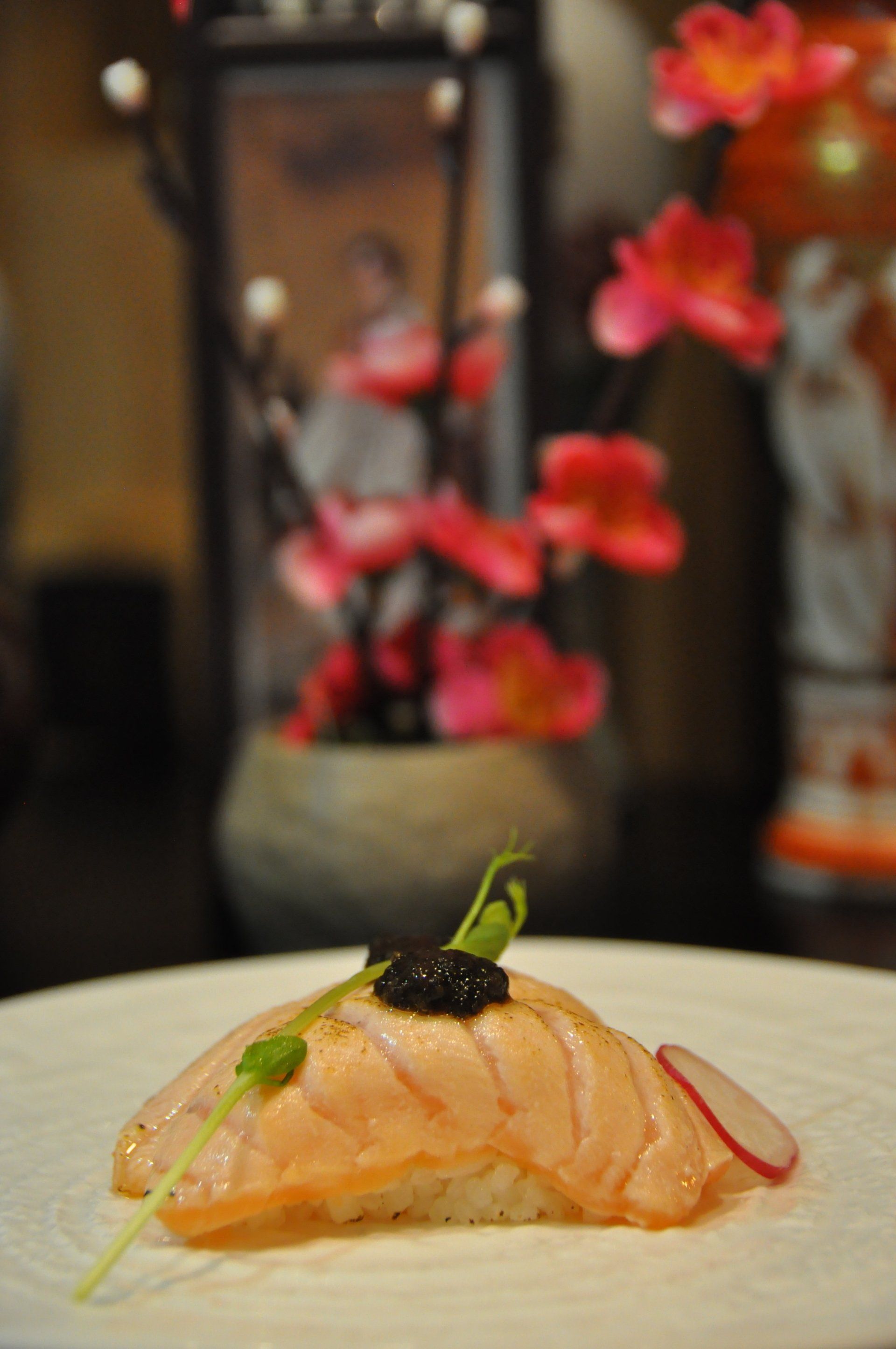 Sushi nigiri su un piatto bianco, guarnito con un elemento decorativo, con fiori sfocati sullo sfondo.