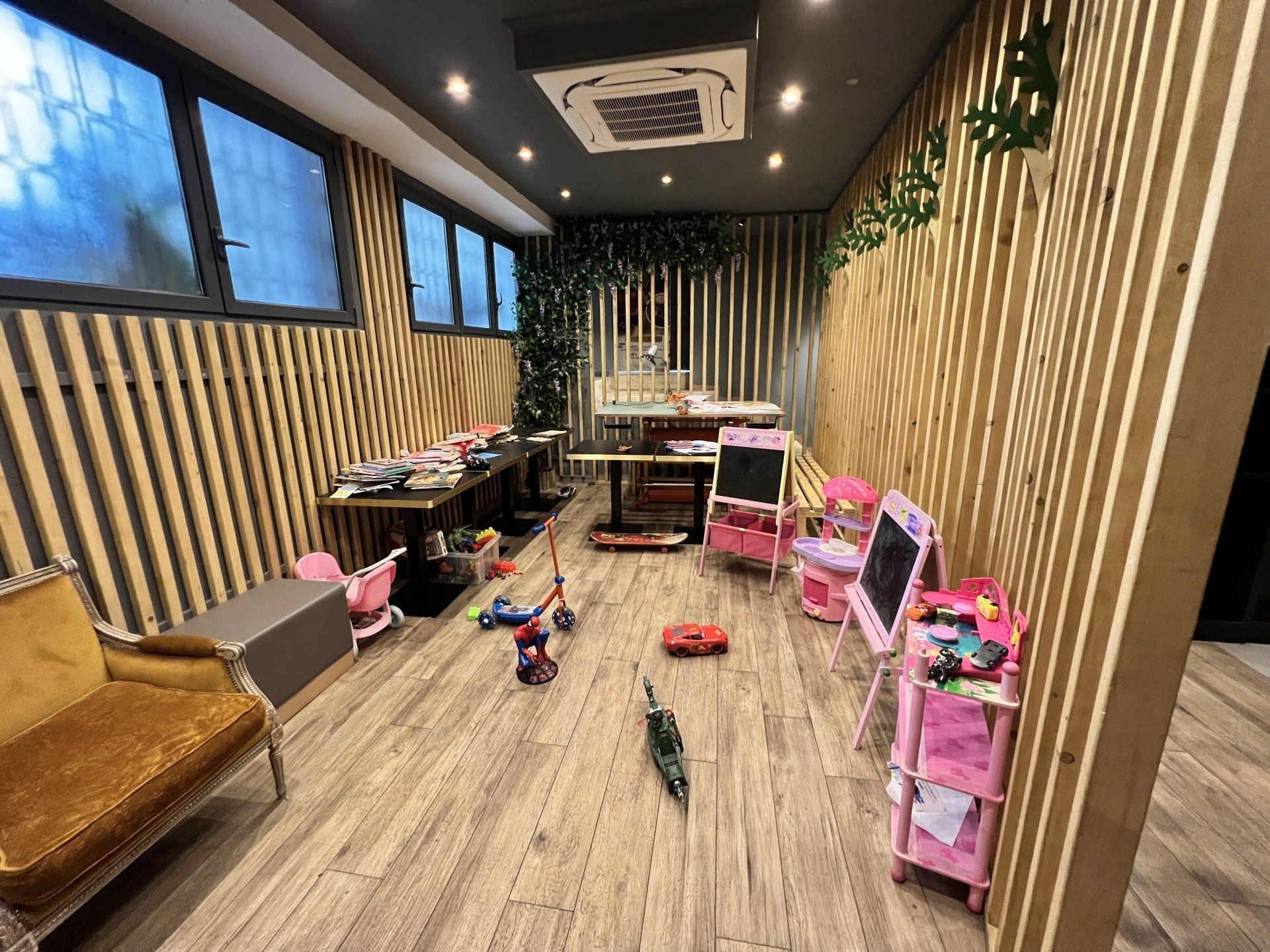 Una piccola stanza dei giochi con giocattoli, carrelli rosa, un divano beige e decorazioni murali a righe sotto luci intense del soffitto.