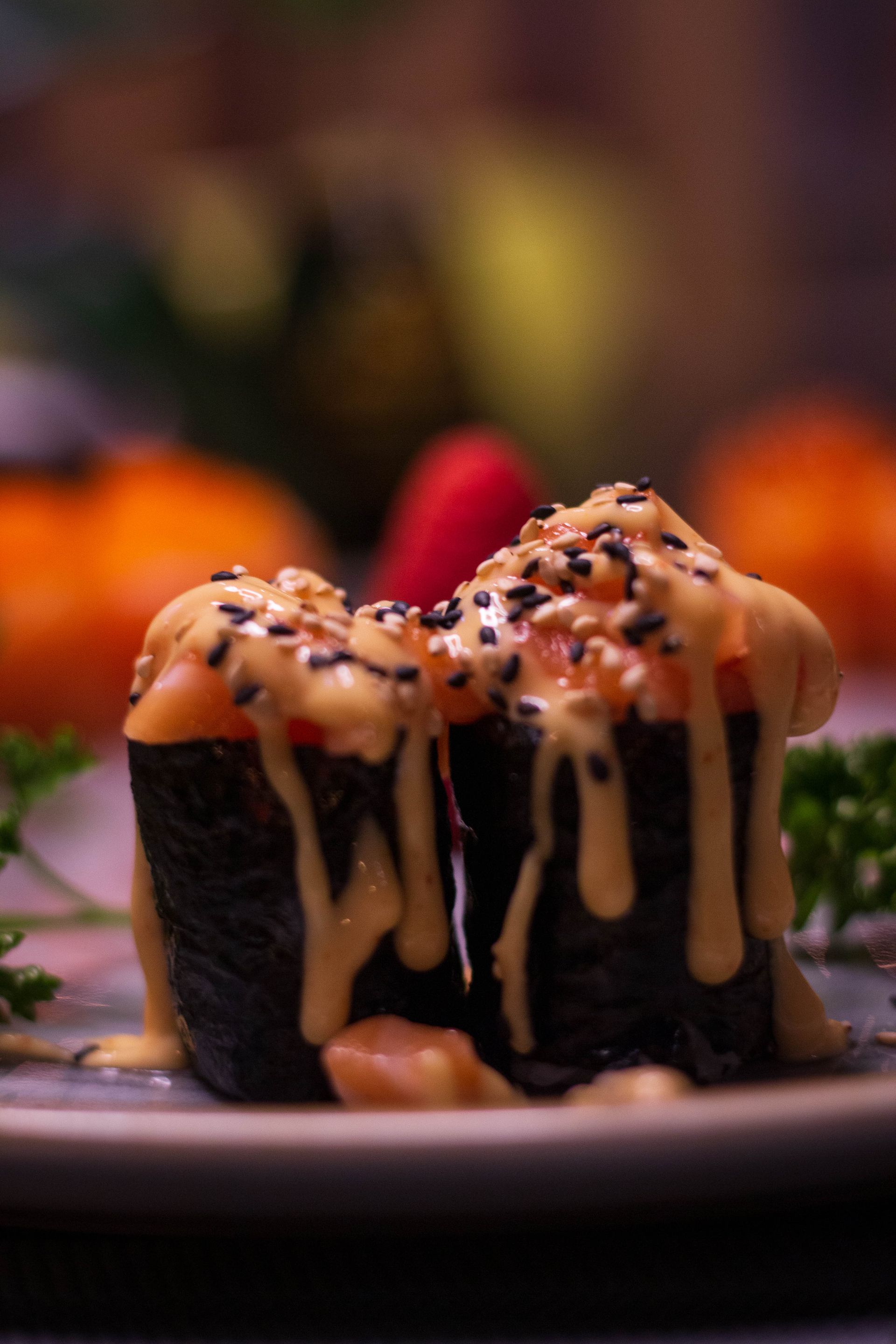 Su un piatto, con uno sfondo sfocato, si possono vedere dei rotolini di sushi conditi con salsa cremosa e semi di sesamo.