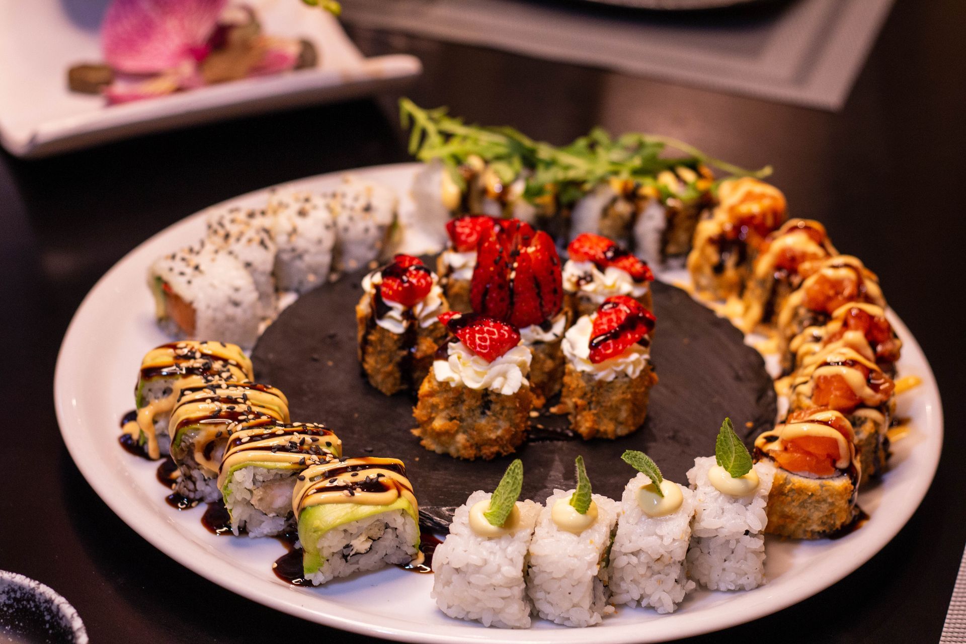 Assortimento di rotolini di sushi disposti in cerchio su un piatto bianco con salsa di accompagnamento al centro.