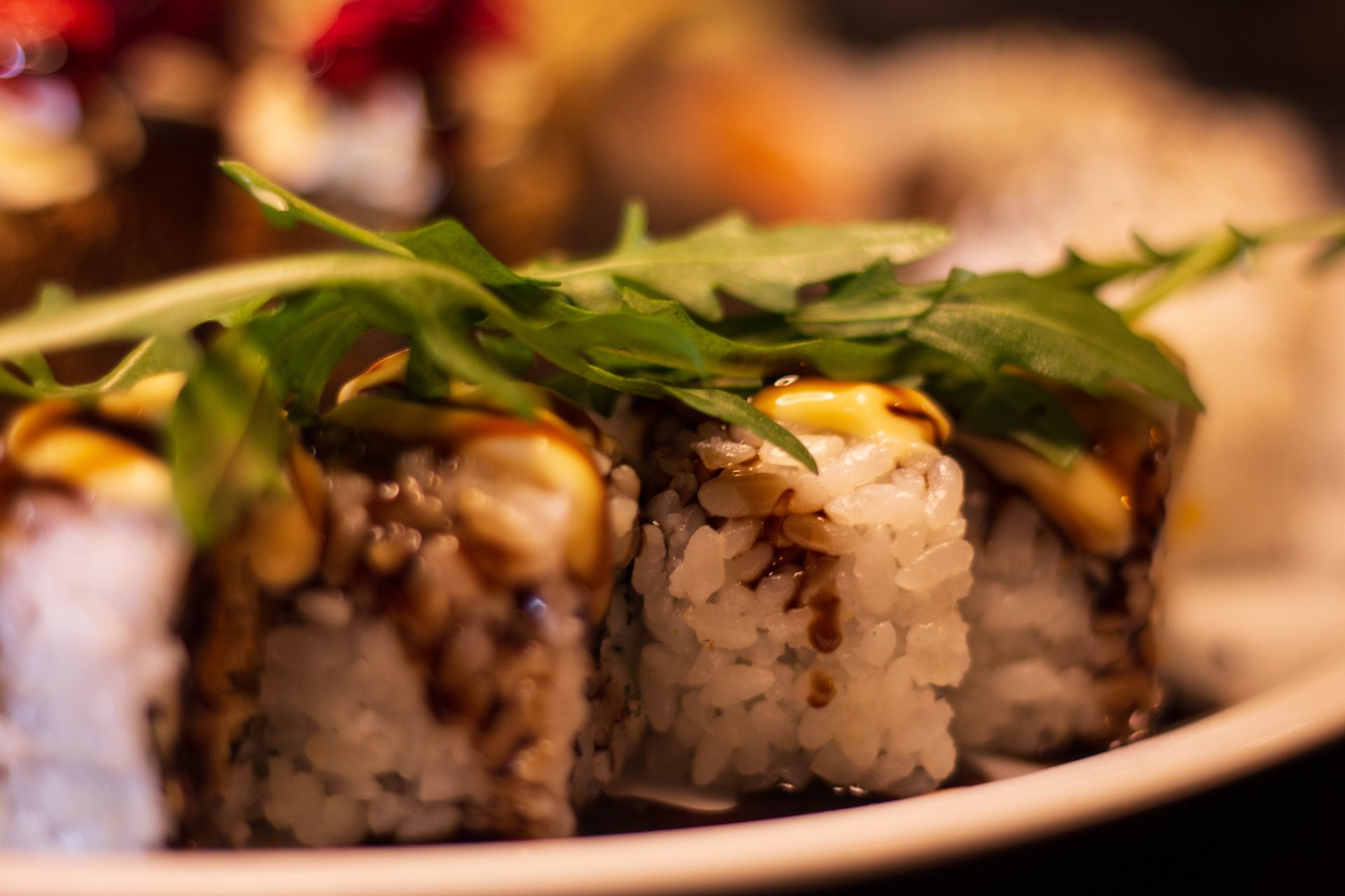 Rotoli di sushi guarniti con verdure e salsa su un piatto bianco illuminato da una luce calda.
