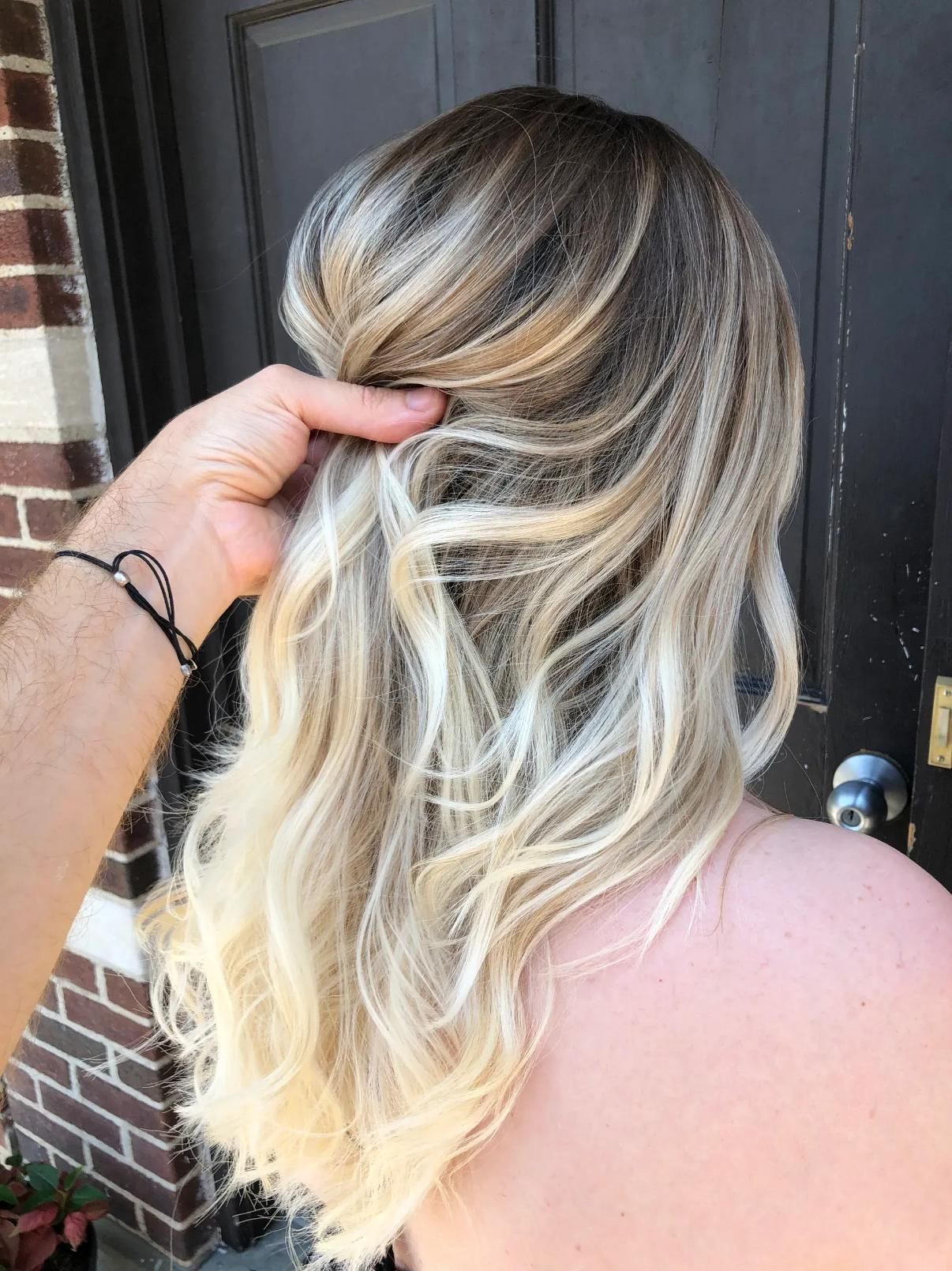 Person's blonde, wavy hair, with darker roots, is held by a hand. Outdoors, near a brick wall and door.