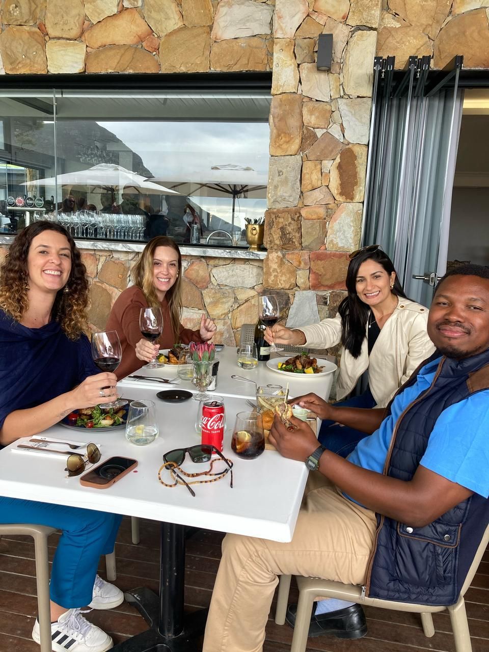Quatro pessoas jantando ao ar livre, sorrindo, segurando bebidas. Mesa branca, parede de pedra ao fundo.
