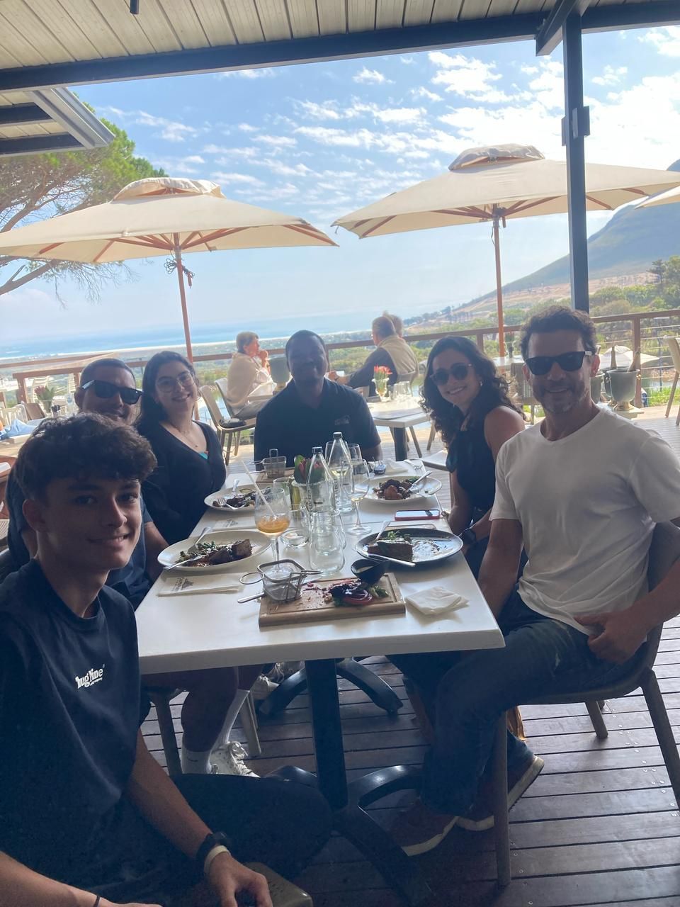 Grupo de pessoas sorrindo em uma mesa ao ar livre, apreciando a comida. Vista para o mar em um dia ensolarado.