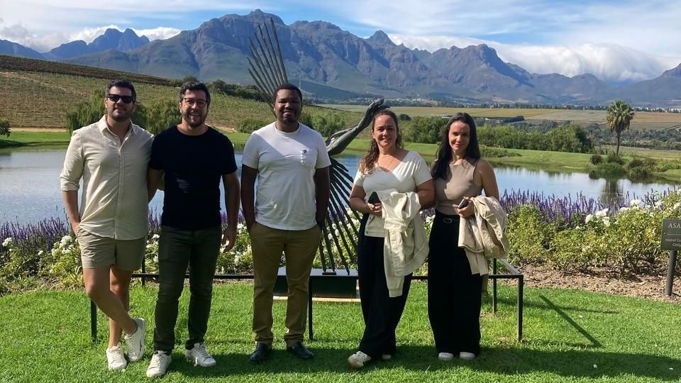 Cinco pessoas posam em frente a um lago, uma escultura e montanhas. Dia ensolarado.