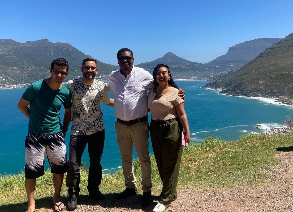 Quatro pessoas sorrindo, posando em uma baía azul com montanhas ao fundo.