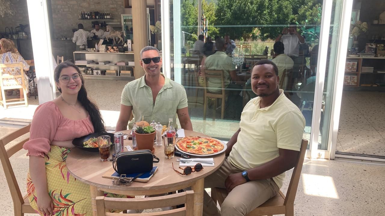Três pessoas sentam-se ao redor de uma mesa em um restaurante ao ar livre. Elas sorriem, com comida na mesa.