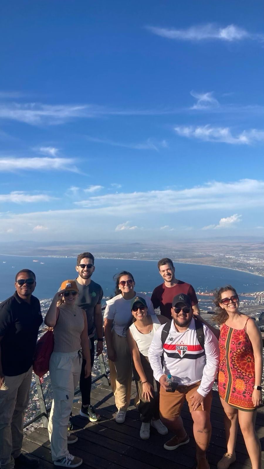 Grupo de pessoas sorrindo para a câmera no pico da montanha com vista para o mar e a cidade.