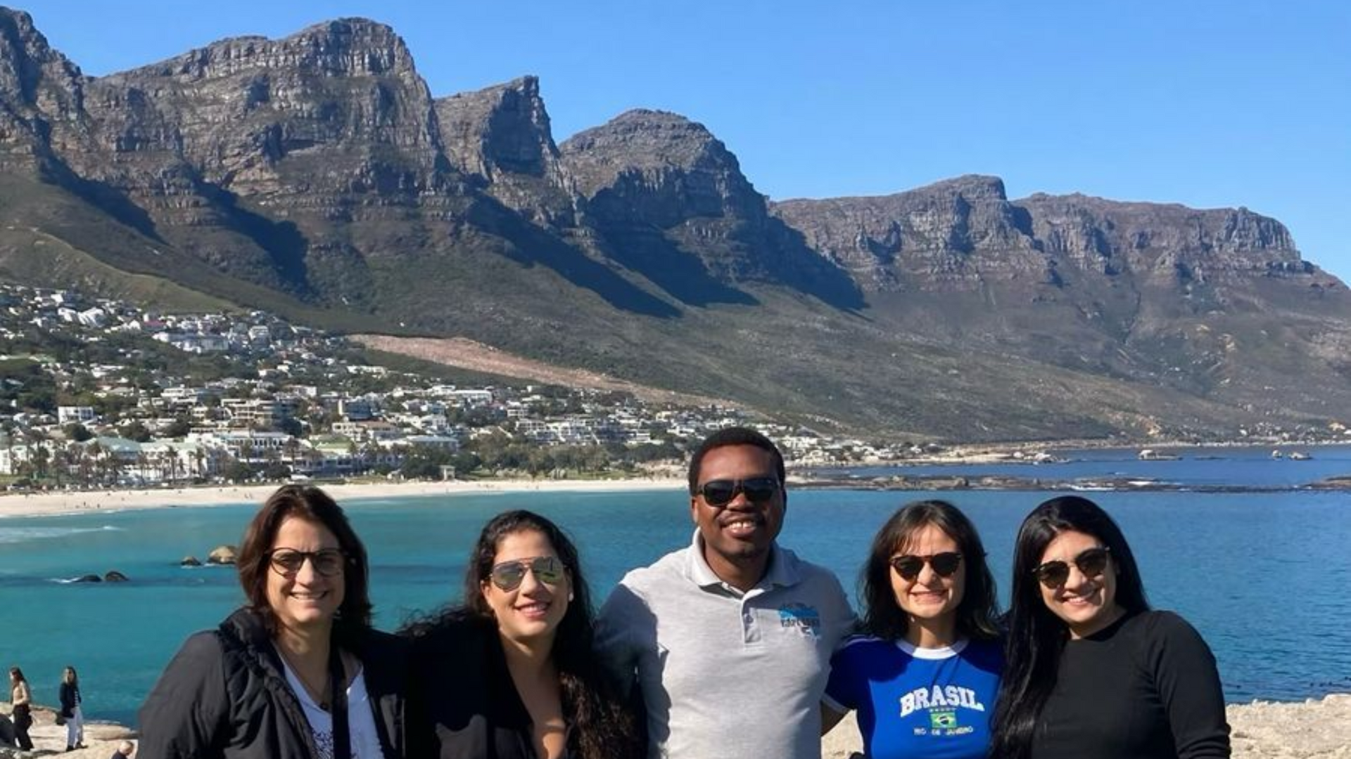 Cinco pessoas sorrindo em frente a uma praia, oceano azul e cadeia de montanhas em um dia ensolarado.
