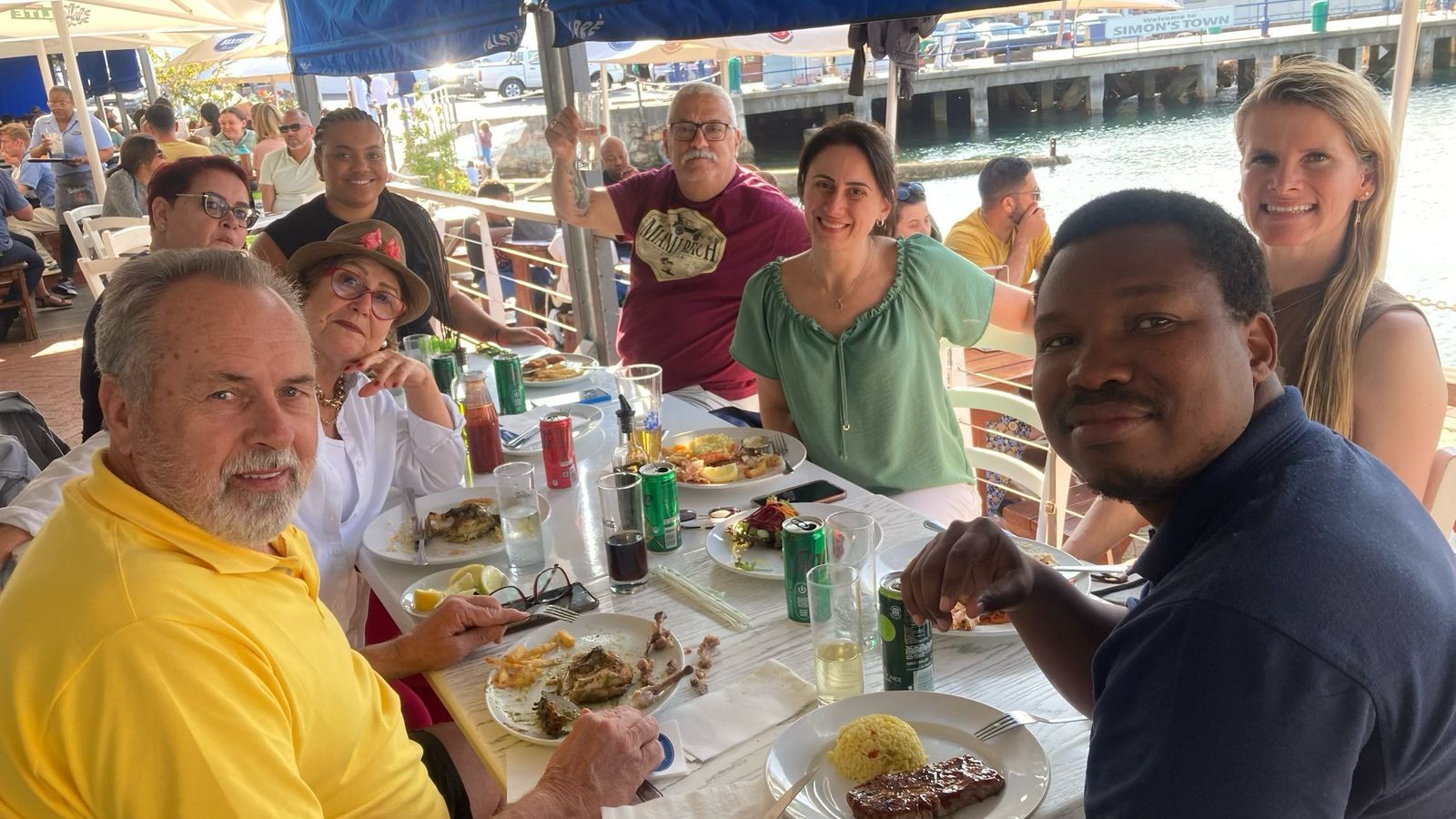 Grupo de pessoas em um restaurante à beira-mar, sorrindo, comendo. Dia ensolarado com água azul ao fundo.