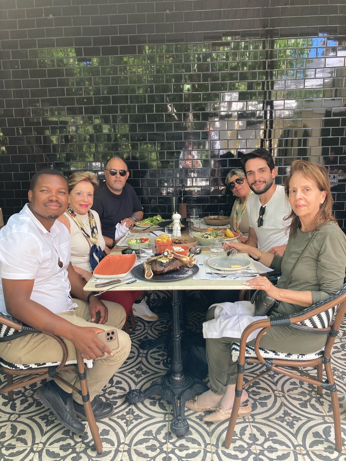 Grupo de pessoas em uma mesa ao ar livre, comendo e sorrindo. Parede de azulejos pretos ao fundo.