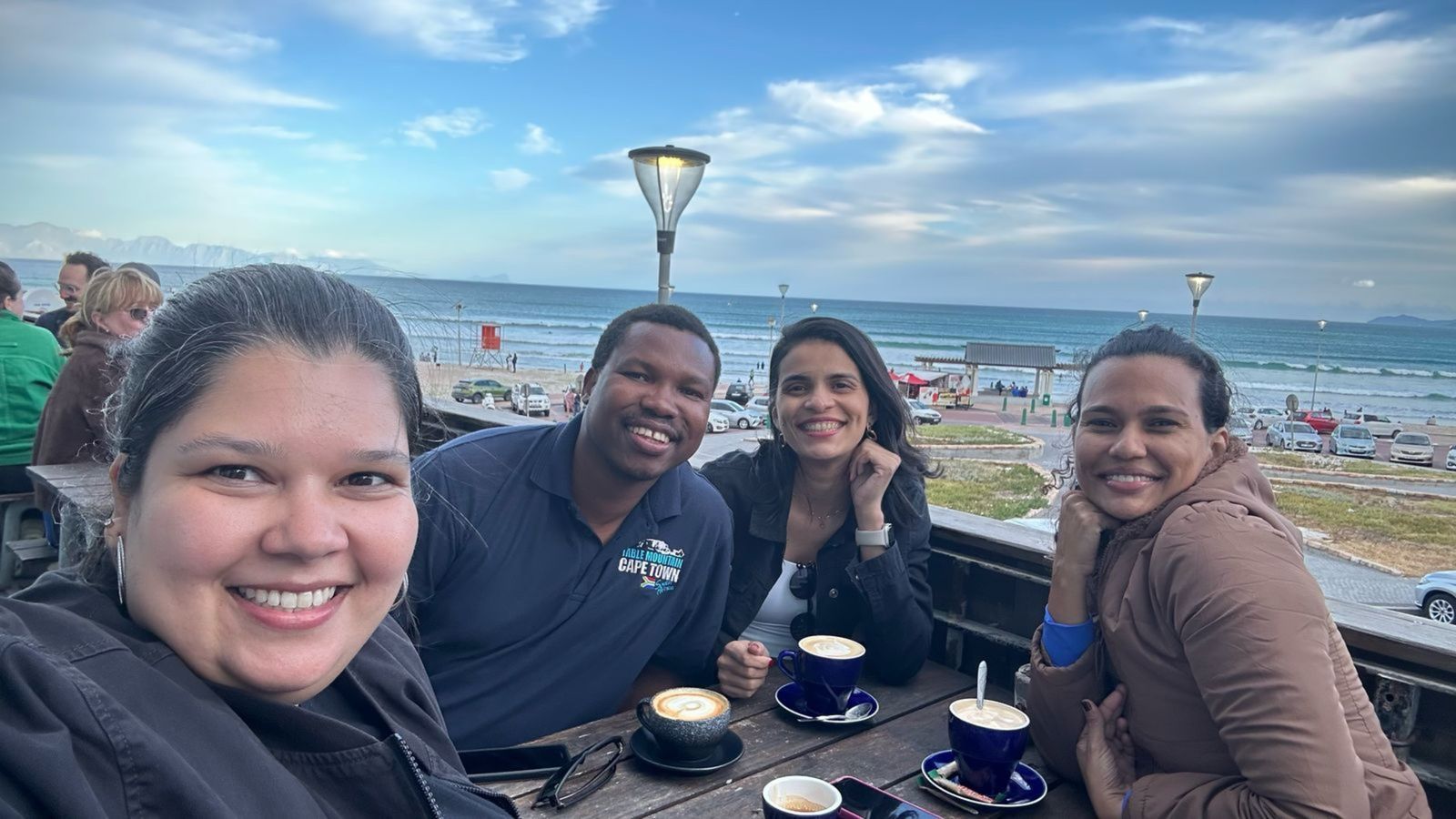 Quatro pessoas sorrindo em uma mesa com café, praia ao fundo. Céu nublado.