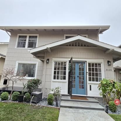 Beige Craftsman house with blue door and open screen, flanked by windows. Grey overcast sky.