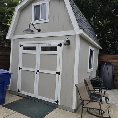 Gray shed with barn-style roof, white trim, black hardware, two lights, two chairs, and a blue trash can.