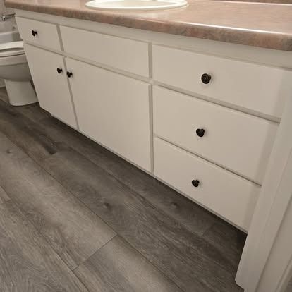 White bathroom vanity with three drawers and two cabinet doors, brown countertop, gray wood-look floor.