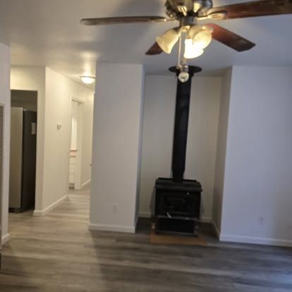 A living room with a wood-burning stove, ceiling fan, and gray flooring. A hallway leads to other rooms.