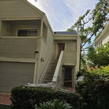 Multi-story house exterior with stairs leading to a doorway, gray siding, garage door, and lush greenery.