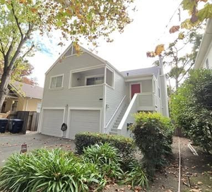 Light gray two-story house with a red front door, stairs, and two garage doors. Lush greenery in front.
