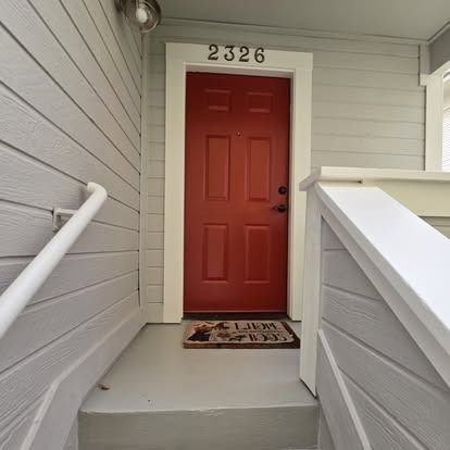 Red door with the number 2326, gray siding, white trim and railing. Welcome mat.