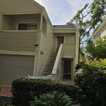 Two-story tan house with a garage and outdoor stairs leading to a second-floor entrance; shrubs in foreground.
