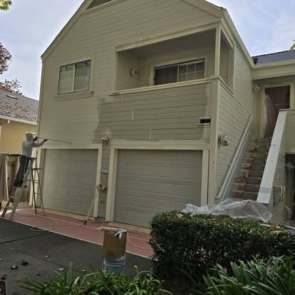 Person painting a two-story beige house with two gray garage doors. The house has a balcony, and a ladder is set up.