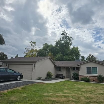 Single-story house with a tan exterior, attached garage, and a car in the driveway. Overcast sky.