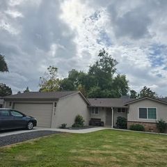 Single-story house with a tan exterior, attached garage, and a car in the driveway. Overcast sky.