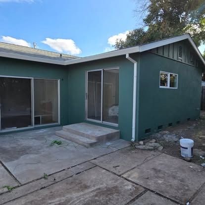 Green house with patio, sliding doors, and small concrete steps under a blue sky.