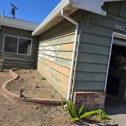 Green house with peeling paint, open garage, and a brick-bordered dirt patch in front.