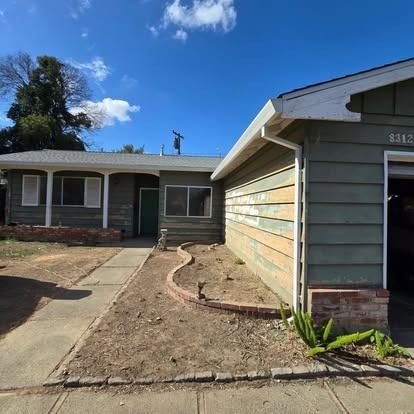 Green house with attached garage under a blue sky. Bare yard, brick edging, and sidewalk.