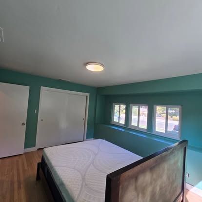 Bedroom with teal walls, wood floor, and large white bed, with closet and windows.