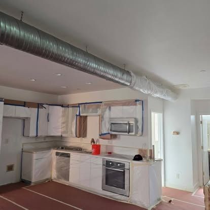 Kitchen under renovation with plastic-covered cabinets and appliances, visible ductwork, and white walls.