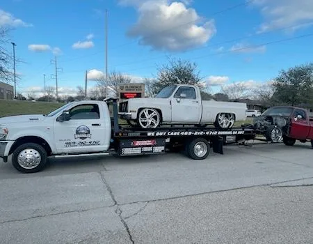 White tow truck hauling a classic white pickup truck and a red pickup truck on a sunny day.