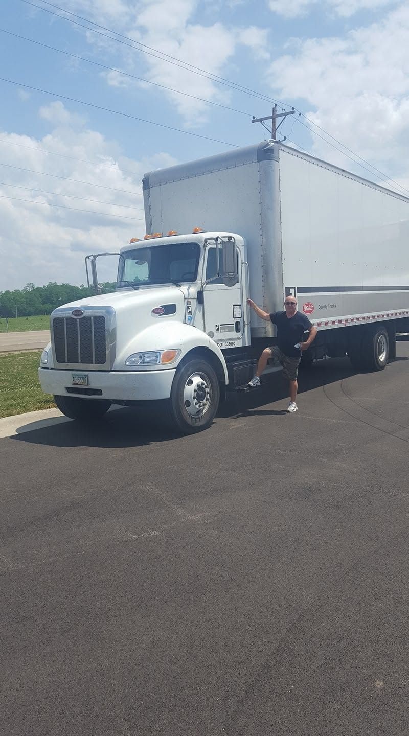 White semi truck with trailer parked on pavement; a person stands beside the cab under a blue sky.