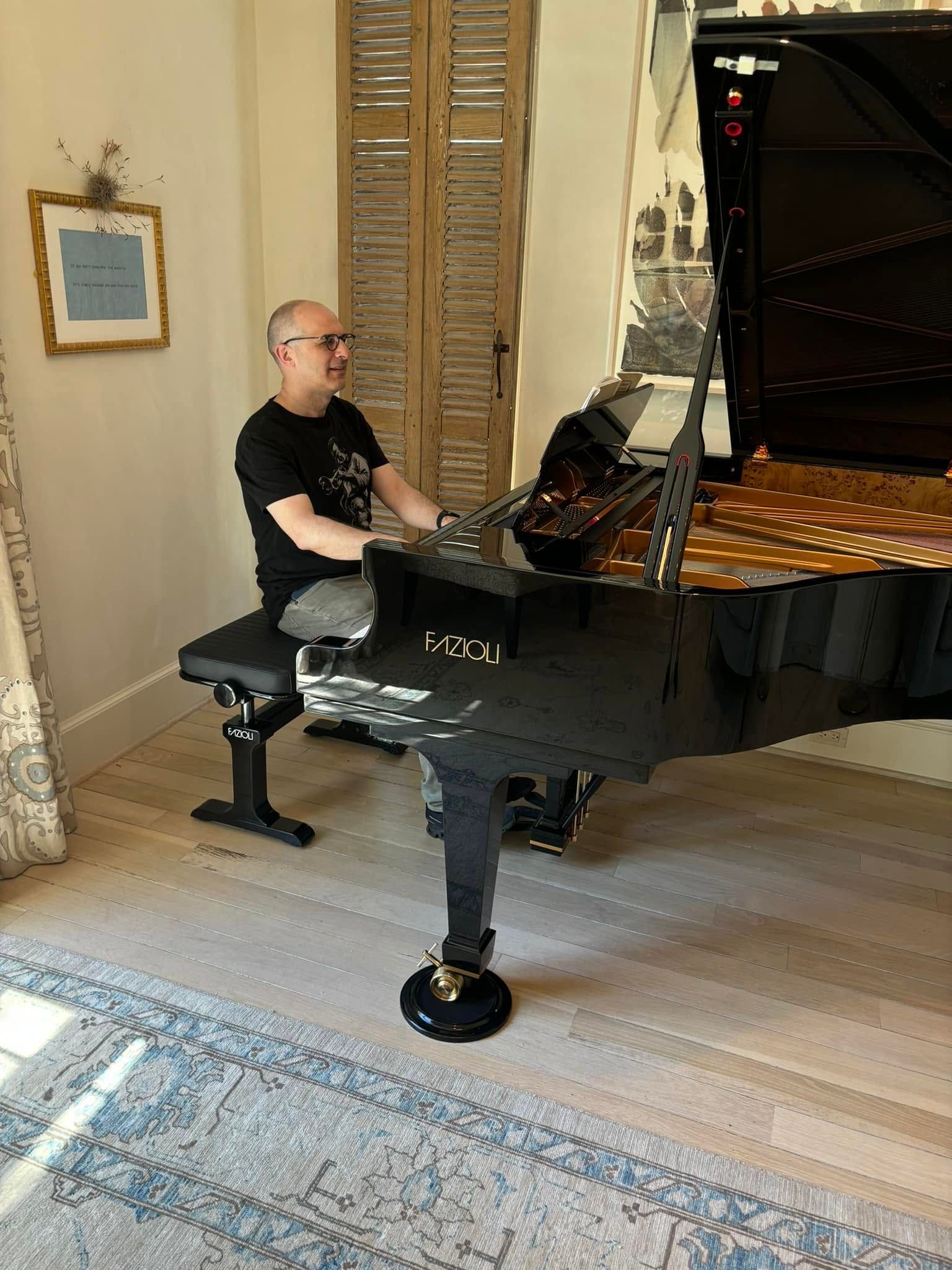 Man seated at a black grand piano in a bright room with a rug and framed wall art.