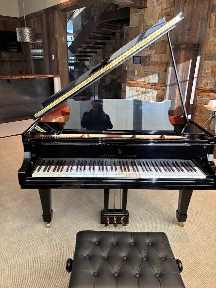 Black grand piano with lid open and tufted bench in a rustic room.