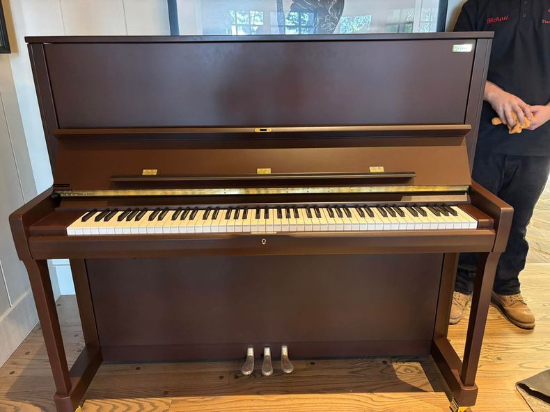 Dark upright piano in a room, with a person standing at the right edge.