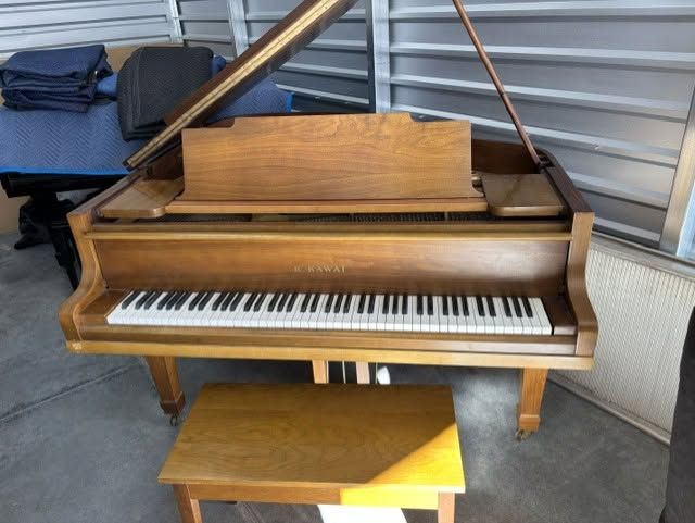 Wooden grand piano with bench in a storage unit, lid open and keyboard visible.