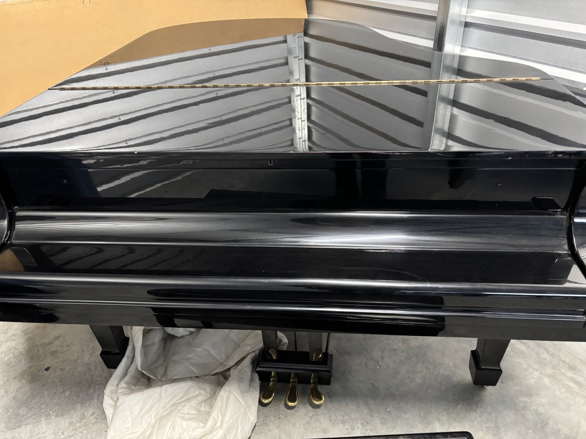 Glossy black grand piano with open lid, shown in a room with a light floor and beige wall.