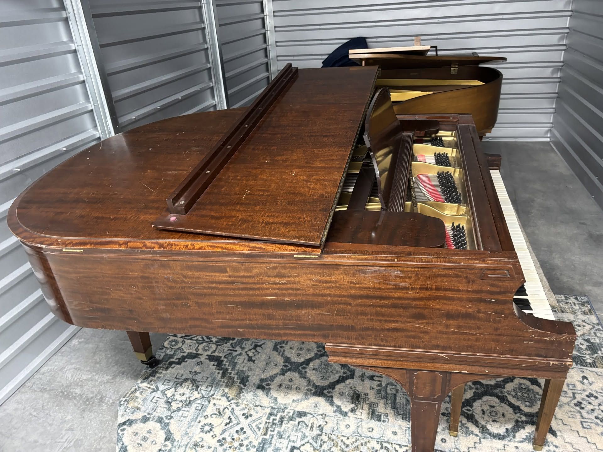 Wooden grand piano with lid open, showing strings and interior, on a patterned rug in a storage space