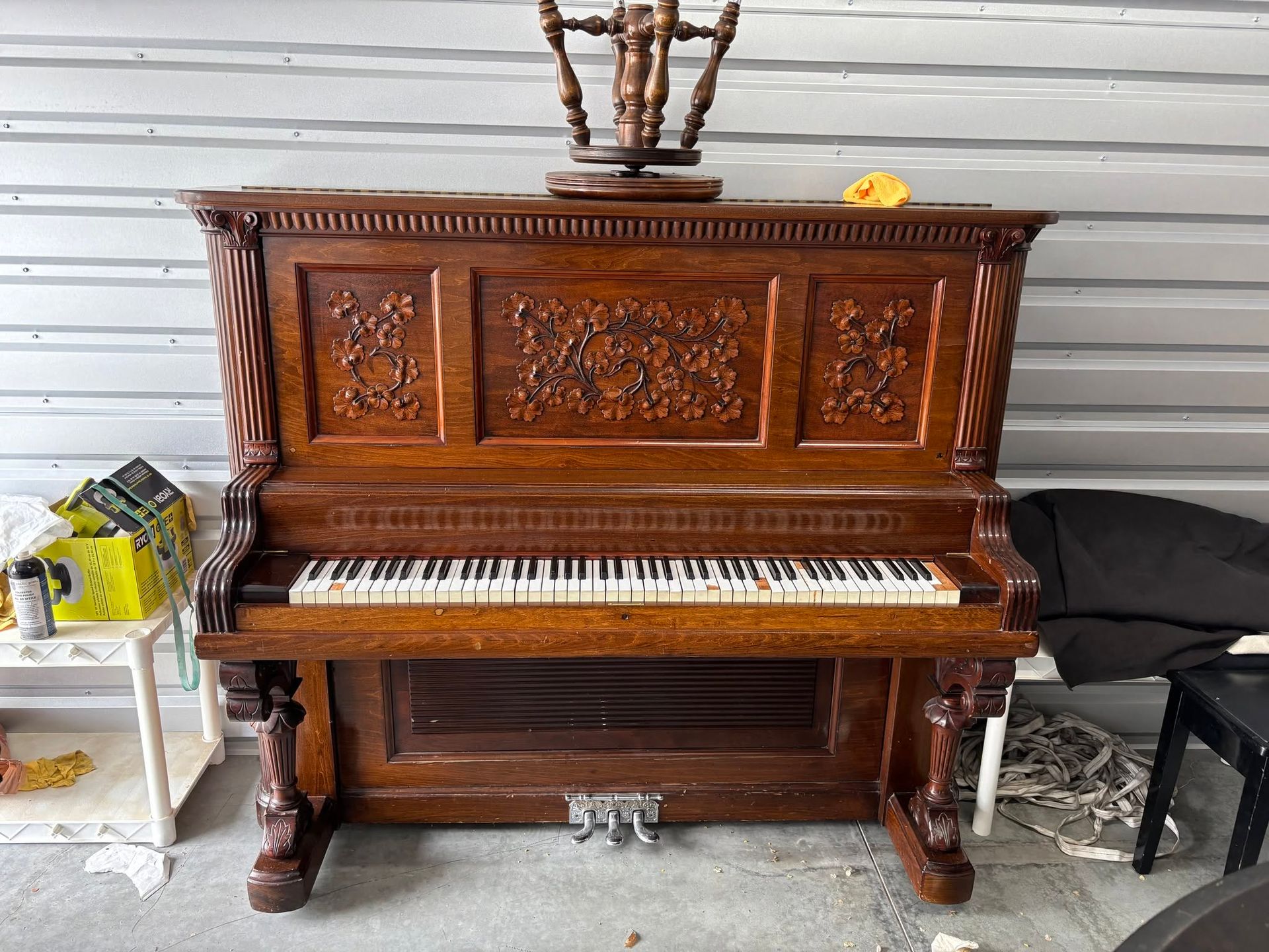 Ornate wooden upright piano in a cluttered garage or storage area, with a yellow cloth on top.