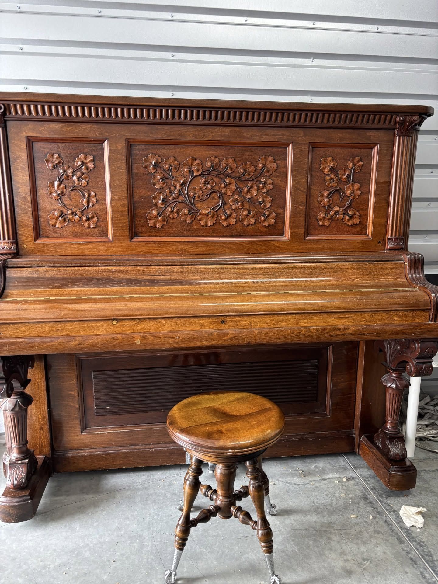 Ornate carved wooden upright piano with a round stool in front, indoors on a concrete floor.