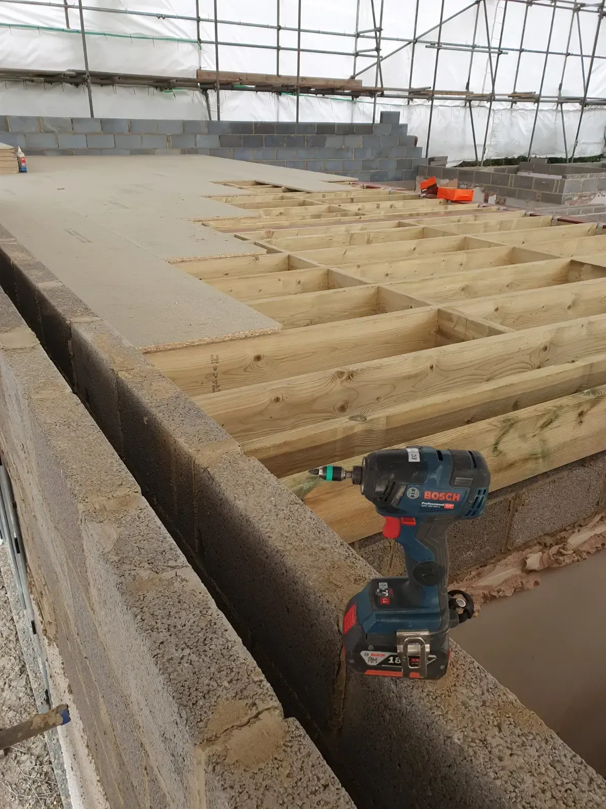 Wooden floor framing under construction with a cordless drill in the foreground and a concrete edge around it